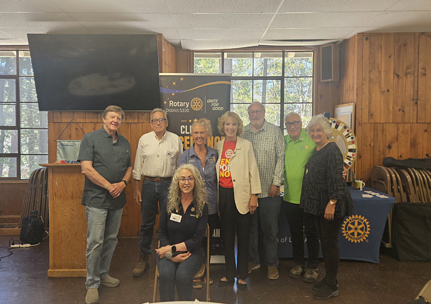 Nine members of the Rotary Club of Lake Arrowhead standing and seated together beside a Rotary banner at a club meeting