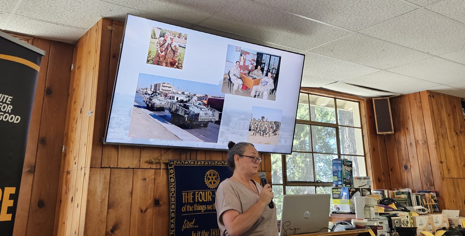 Sage Moritz speaking into a microphone at a Rotary Club of Lake Arrowhead meeting, with a screen behind her showing photographs from her Army deployment to Iraq