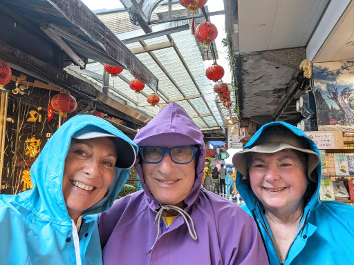 Three Rotarians in brightly colored hooded raincoats smiling on a covered street lined with red lanterns