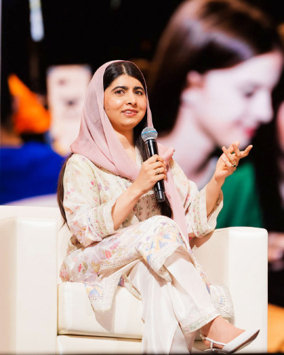 Malala Yousafzai seated on stage speaking into a microphone, with her portrait projected behind her
