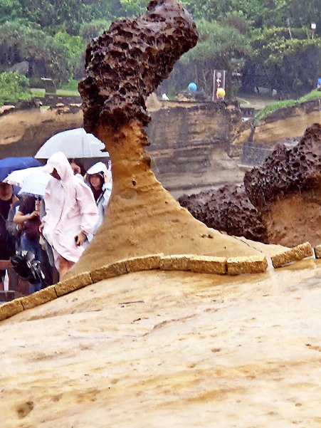 The Queen's Head rock formation at Yehliu Geopark, with visitors in rain gear under umbrellas behind it