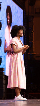 Poet Salome Agbaroji reciting on stage in a pink dress, with her image on the screen behind her